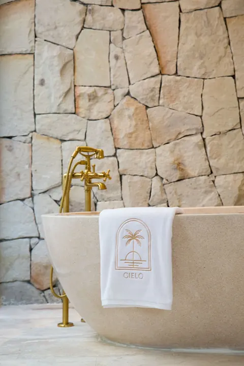 Freestanding stone bathtub with a brass floor-standing mixer against a rough stone wall, a towel embroidered with the Cielo logo over its rim.