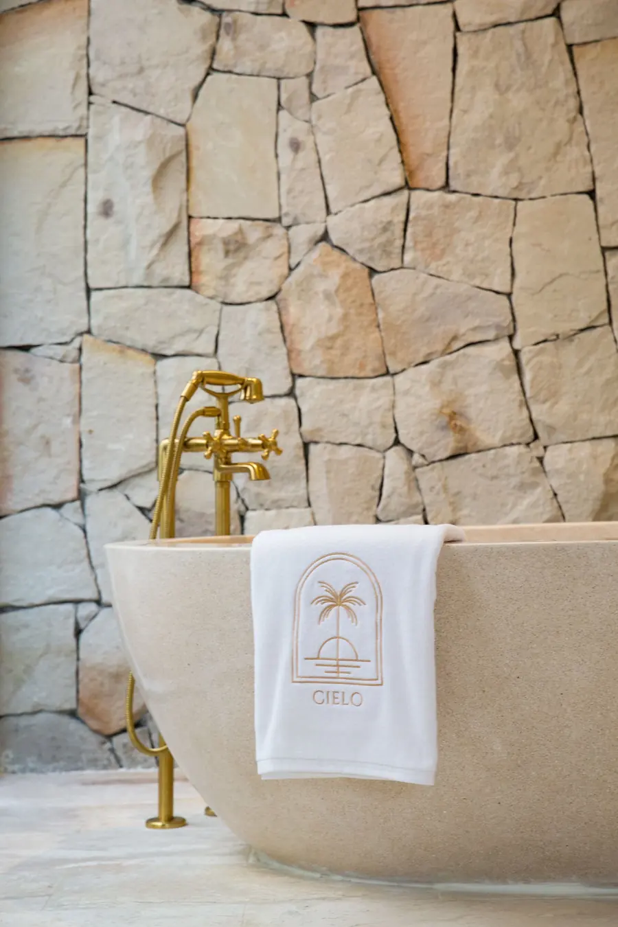 Freestanding stone bathtub with a brass floor-standing mixer against a rough stone wall, a towel embroidered with the Cielo logo over its rim.