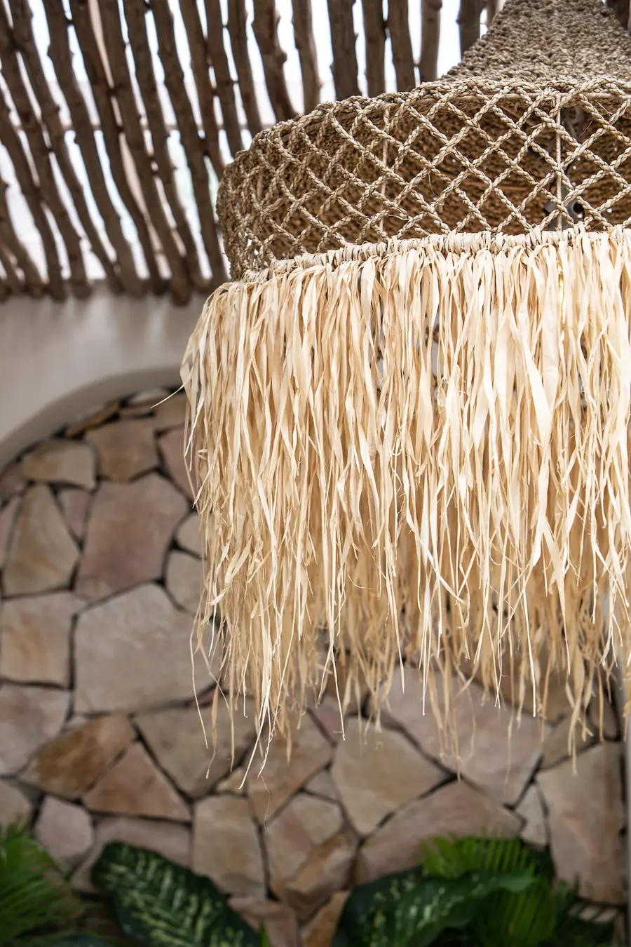 Raffia fringe pendant lamp hanging from a bamboo pergola in front of an arched stone wall with tropical plants.