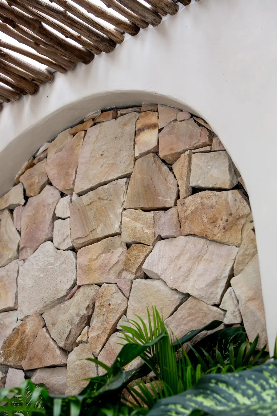 Arched niche of rough pink-and-cream stone under a bamboo pergola, with tropical plants at its base.