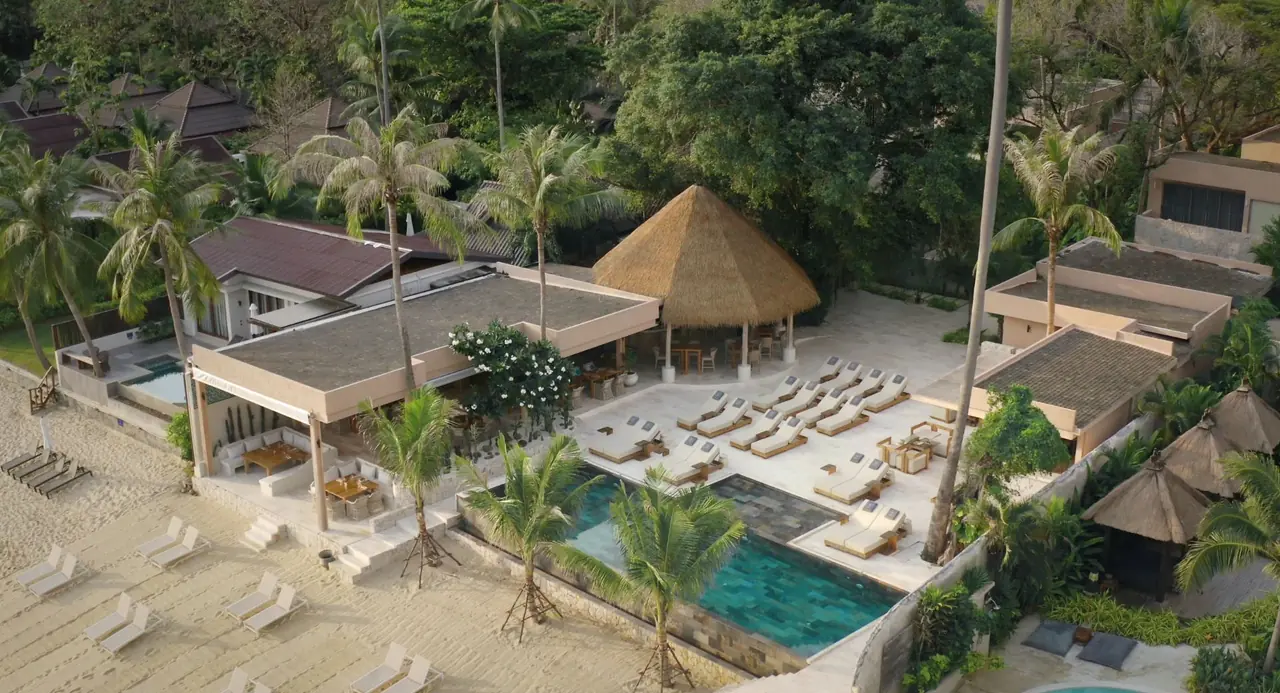 Aerial view of the beachfront: an infinity pool lined with sun loungers, an open-sided restaurant and a thatched sala under palm trees, with white loungers on the sand below.