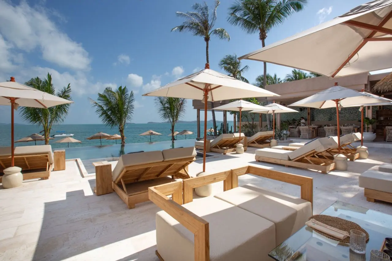 Teak sun loungers and white parasols around an infinity pool at the edge of the sea, with palm trees and distant hills across the water.