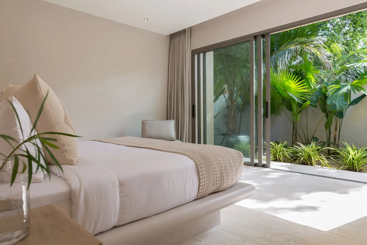 A bed with a woven throw facing open sliding glass doors onto a sunlit private garden of palms.