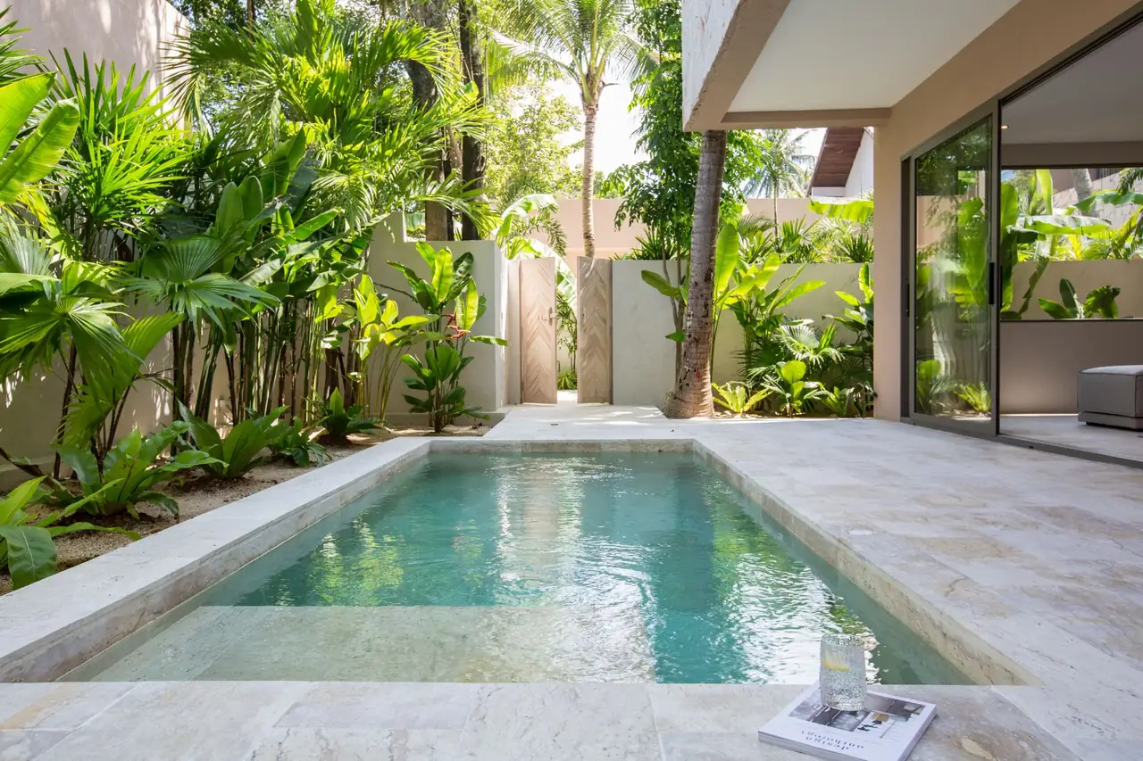 A private plunge pool in a travertine courtyard lined with palms and banana plants, with a glass of water on a magazine at the pool edge.