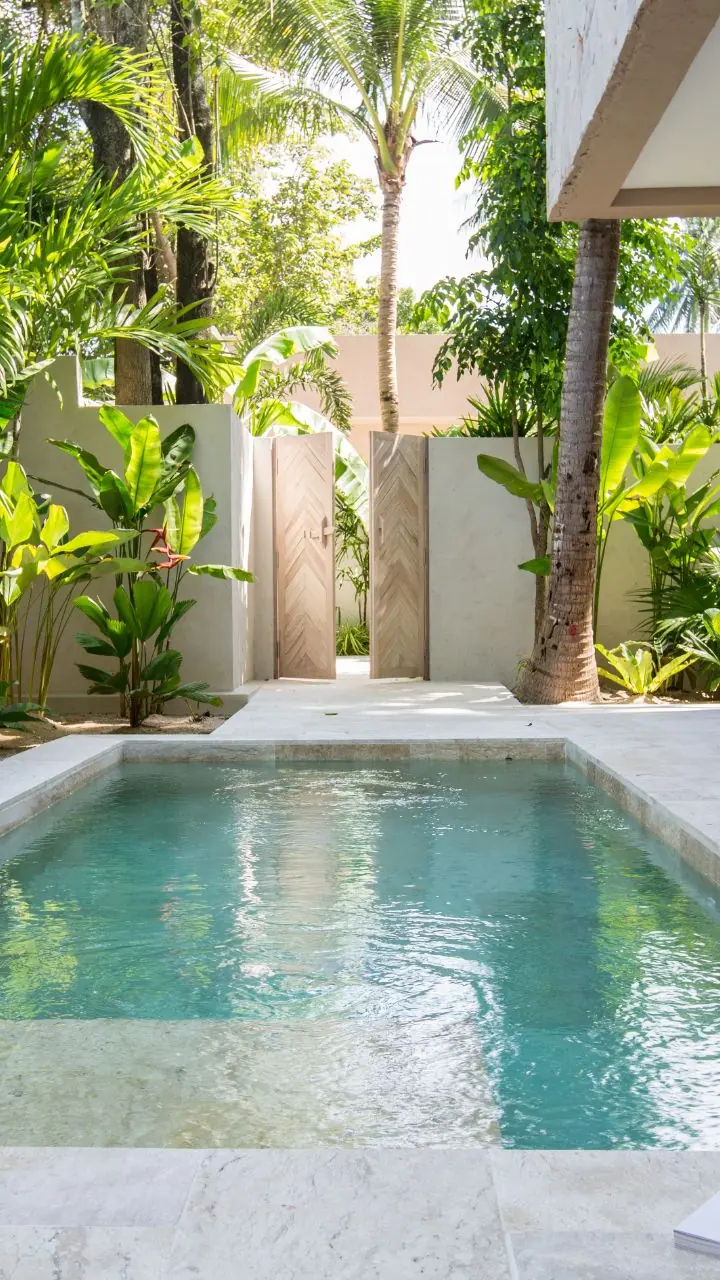 A private plunge pool in a travertine courtyard lined with palms and banana plants, with a glass of water on a magazine at the pool edge.