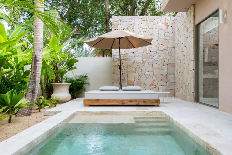 A private plunge pool in a walled courtyard with a cushioned daybed under a parasol, a rough stone wall and tropical plants.