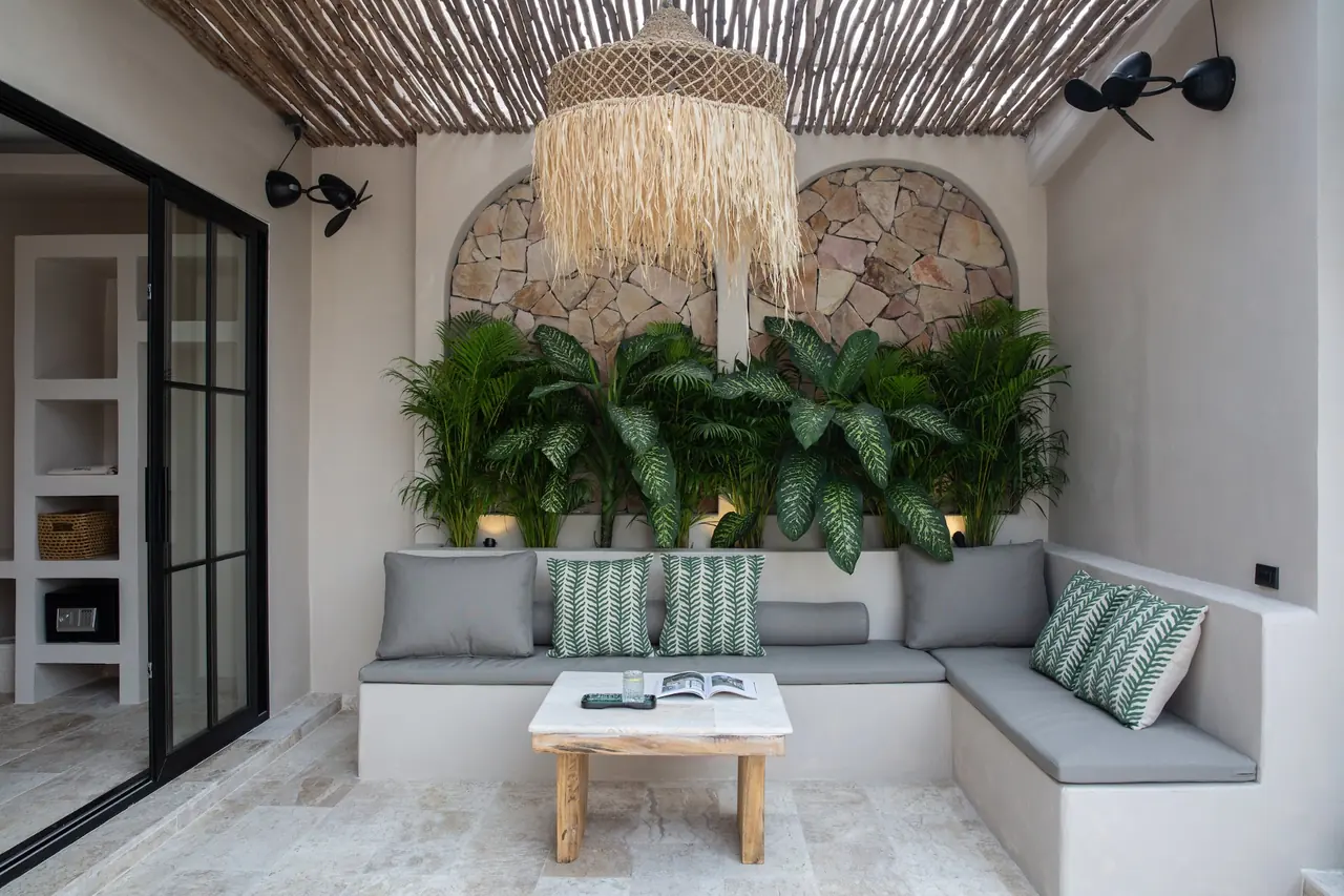 A covered lounge with a raffia pendant lamp under a bamboo pergola, built-in grey sofas with green leaf cushions and two stone arches planted with tropical plants.