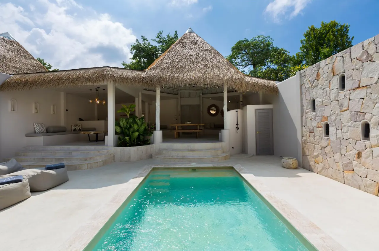 A turquoise pool leading to the open thatched sala of a pool villa, with a stone feature wall and trees under a blue sky.