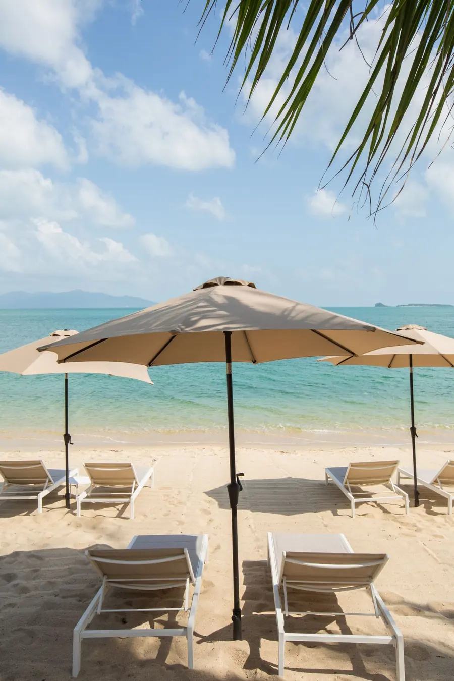 White sun loungers under beige parasols on the sand, with a turquoise sea, distant islands and a palm frond overhead.
