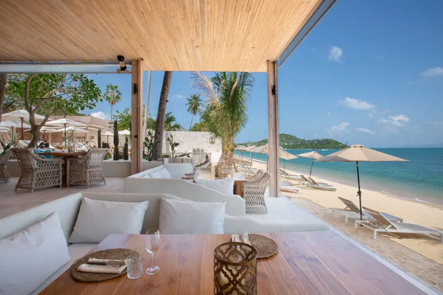 KohCo Beach restaurant under a timber ceiling: a set table and white cushioned banquettes open onto the beach, with parasols, sun loungers, palms and the sea.