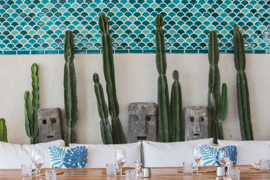 Tall cacti and carved stone face masks along a white banquette with blue leaf-print cushions, under a band of teal fish-scale tiles, with set tables in front.