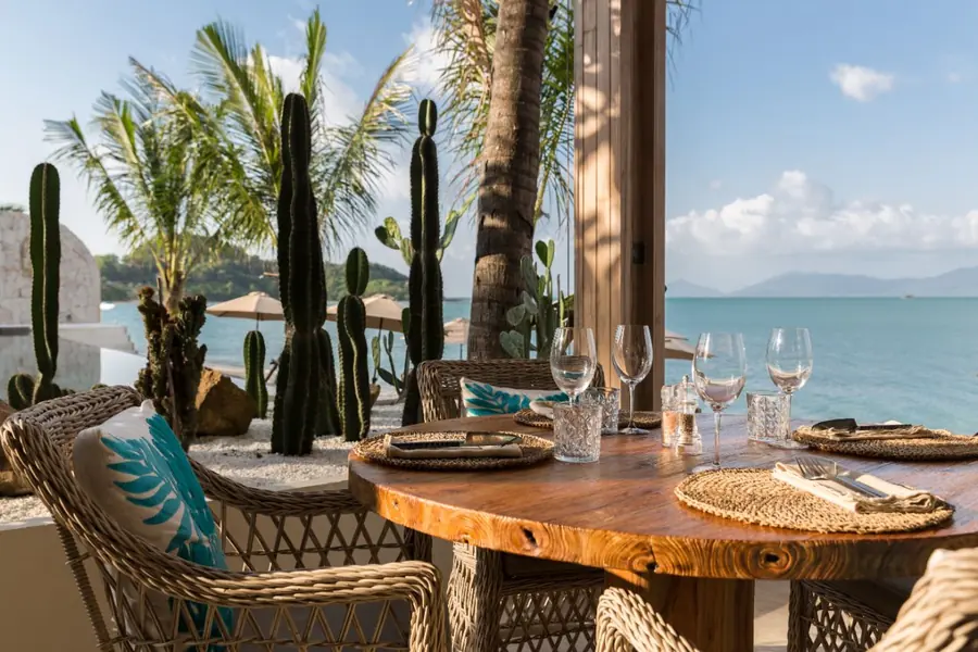 Round teak table set with glasses and rattan placemats, wicker chairs with a teal leaf cushion, tall cacti and palms, and the sea and hills beyond.
