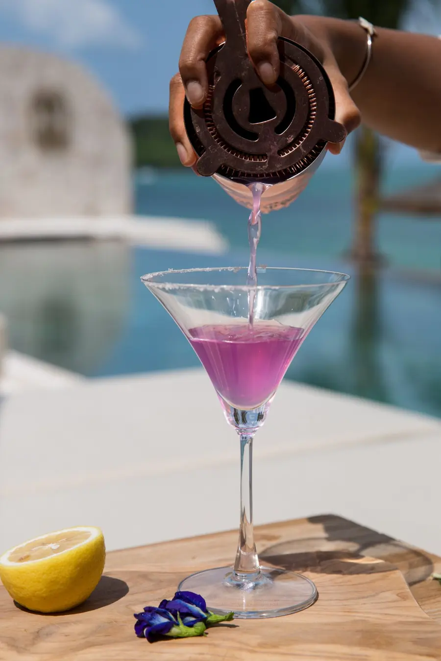 A bartender's hand straining a purple cocktail into a salt-rimmed martini glass on a wooden board with half a lemon and a blue pea flower, with a pool and the sea blurred behind.
