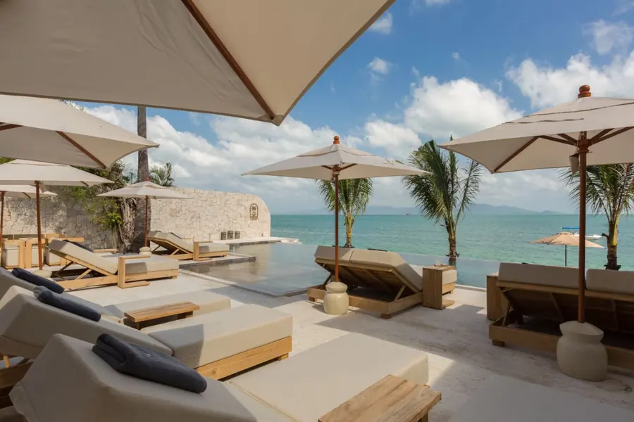 Sun loungers with navy towels under white parasols beside the infinity pool, with the sea, palms and hills on the horizon.