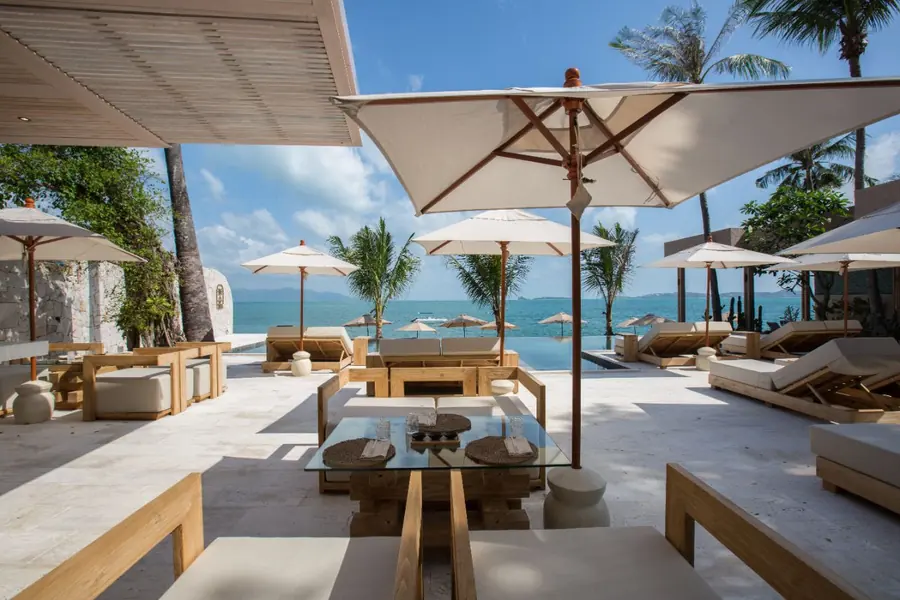 Teak lounge sofas around a low glass table set with glasses and placemats under a white parasol, beside the infinity pool with the sea and palms behind.