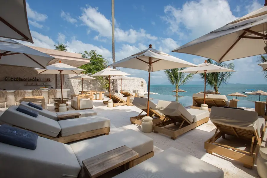 Cushioned loungers and parasols on the pool deck, with the bar and its shelf of bottles at left and the infinity pool and the sea at right.