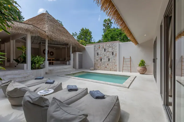Grey bean-bag loungers with folded towels beside the villa pool, with the thatched open sala, a stone feature wall, a bamboo ladder and a terracotta pot.