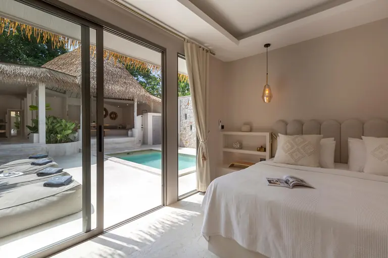 Bed with a scalloped headboard beside full-height glass doors open to the turquoise pool, loungers and the thatched sala.