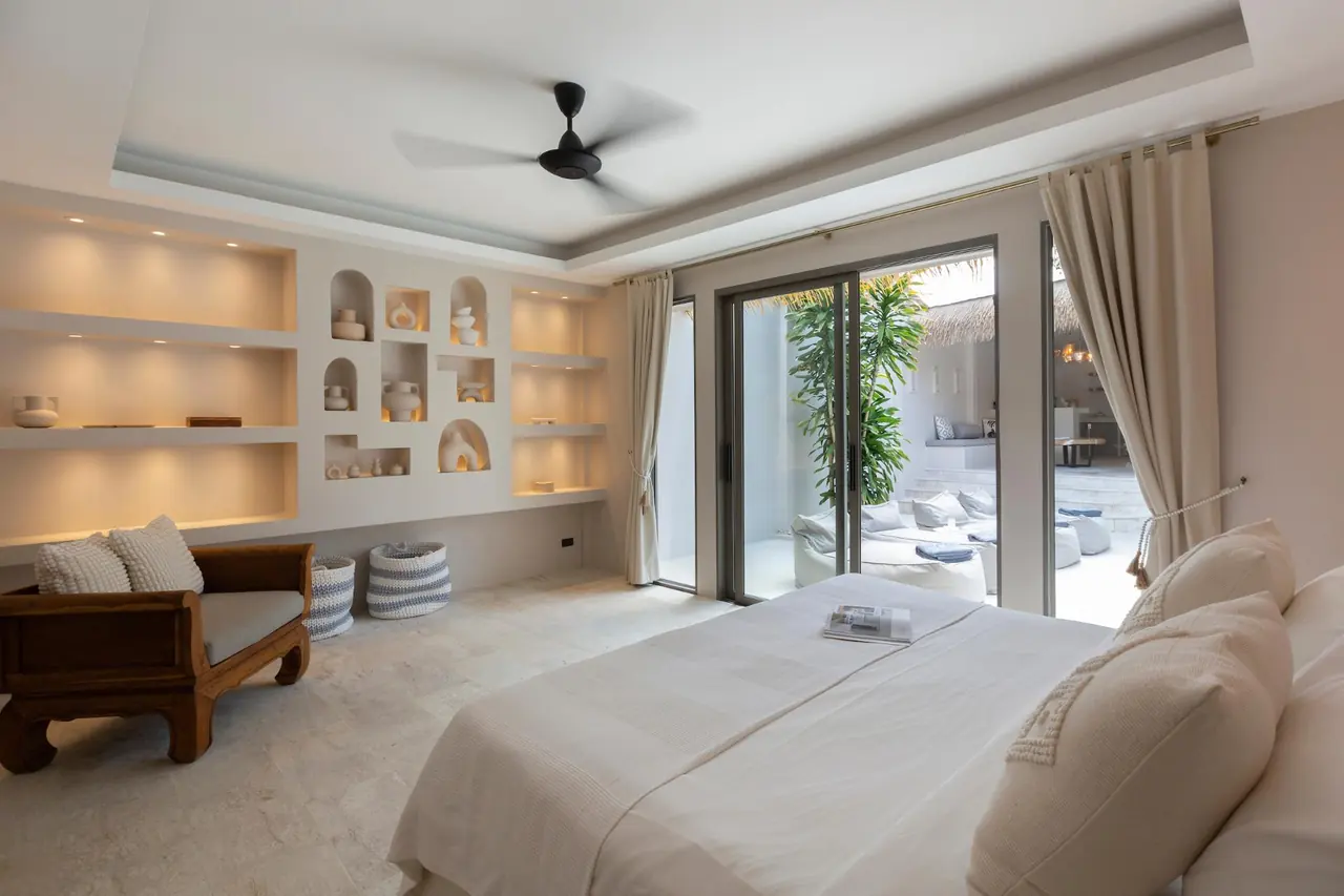 Bedroom with a wall of lit shelves and arched niches, a carved wooden daybed, a ceiling fan and glass doors to the pool courtyard.