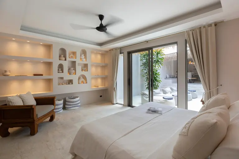 Bedroom with a wall of lit shelves and arched niches, a carved wooden daybed, a ceiling fan and glass doors to the pool courtyard.