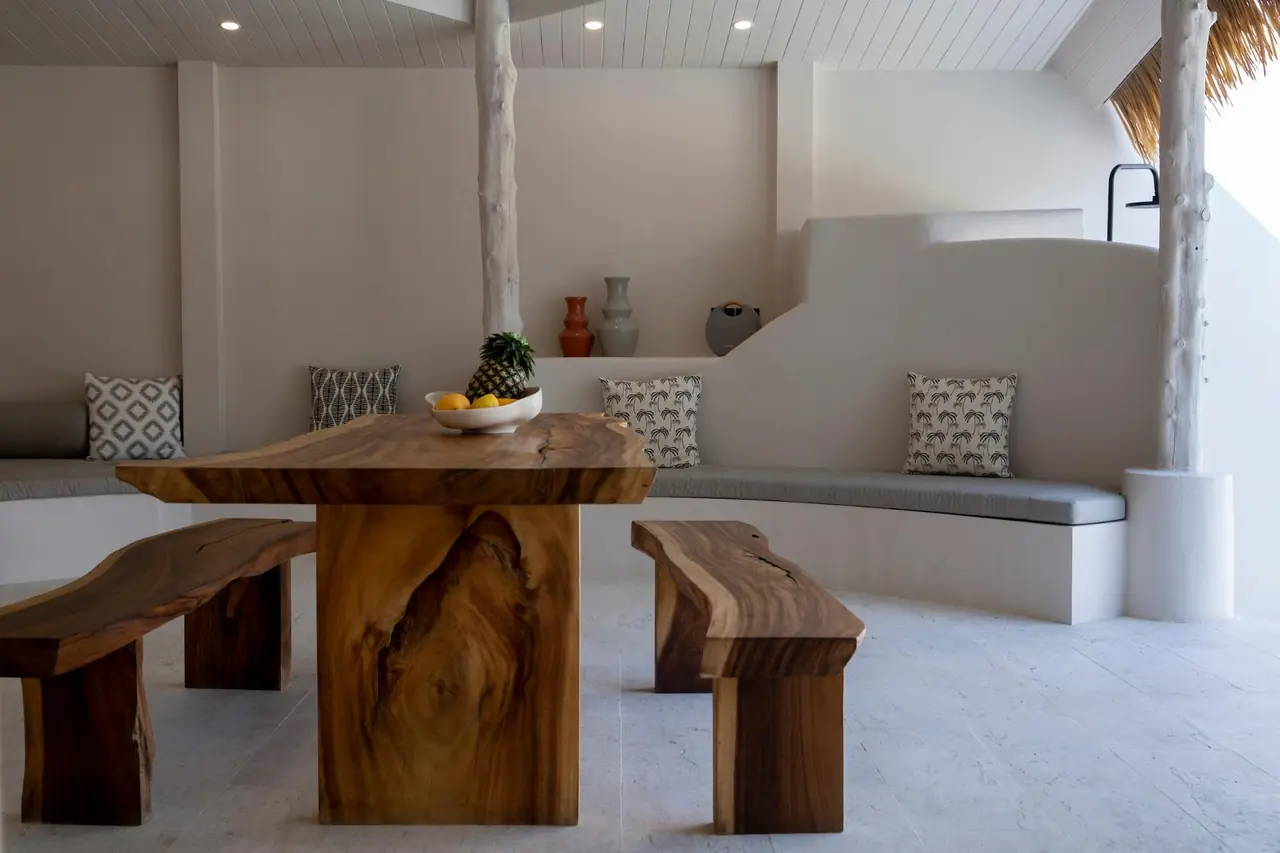 Live-edge teak dining table and benches with a bowl of fruit and a pineapple, in front of a curved built-in sofa with patterned cushions.