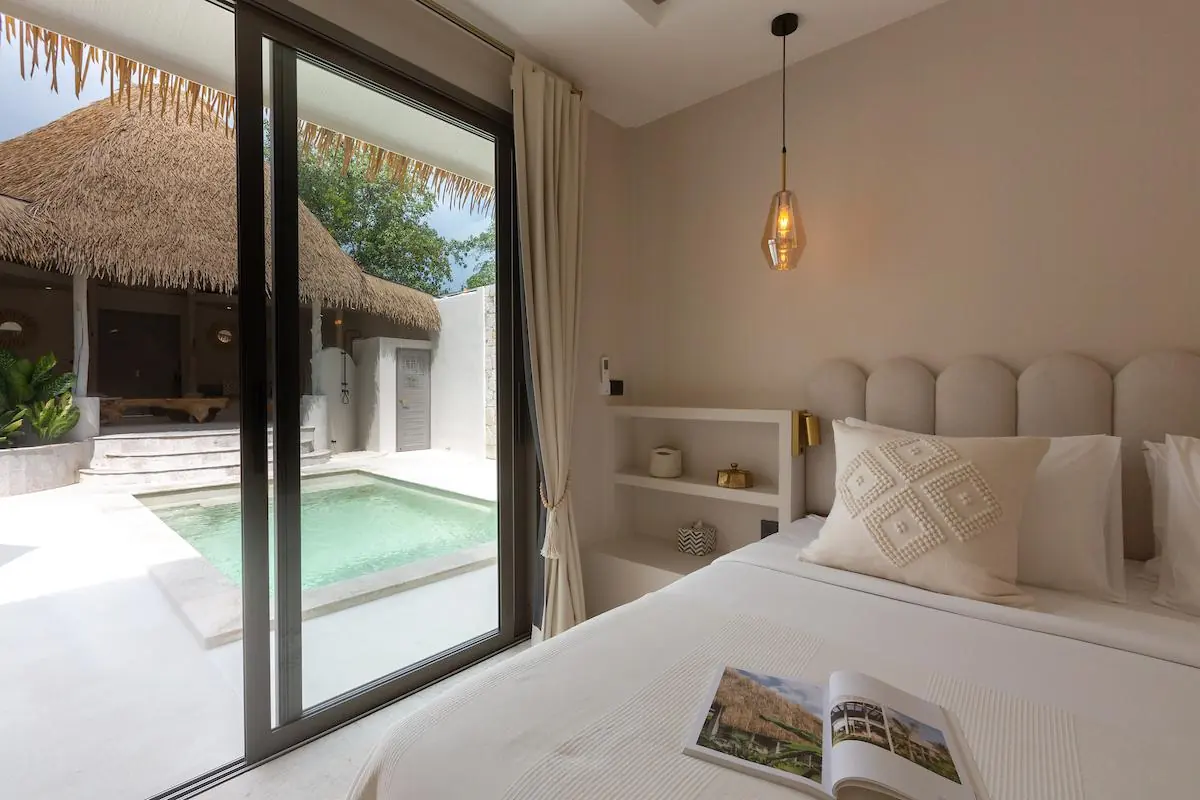 Bedroom with a sliding glass door open onto the plunge pool and thatched sala, and an open magazine on the bed.