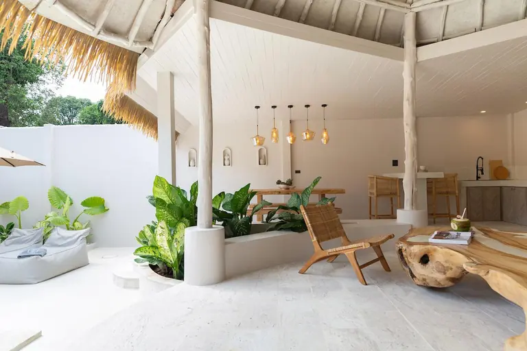 Open-sided thatched sala with a teak-root coffee table, a rattan lounge chair, a dining table under amber pendant lamps and potted tropical plants.