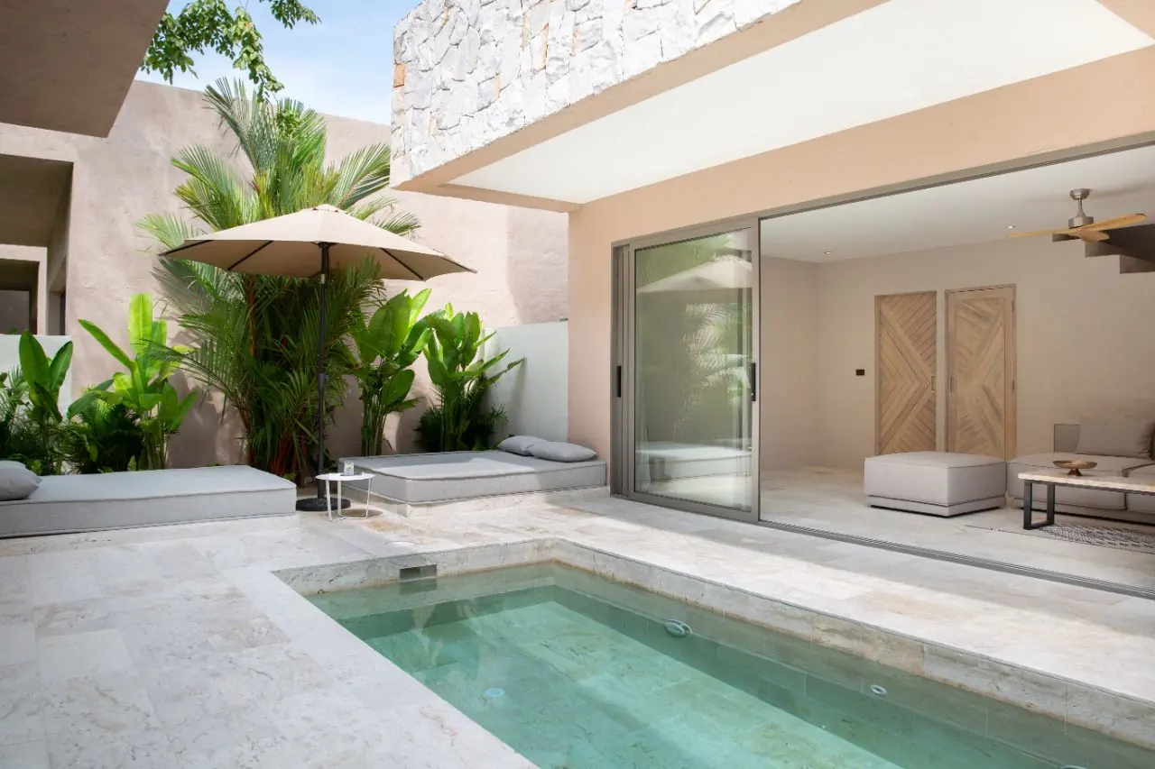 Courtyard plunge pool with cushioned daybeds under a parasol and tropical plants, beside the glass-fronted living room.