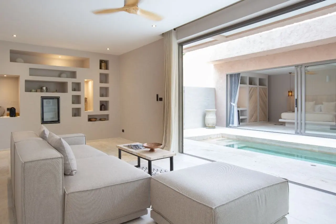 Living room with a grey sofa and ottoman and lit wall niches, its wide glass door open to the plunge pool and a bedroom across the courtyard.