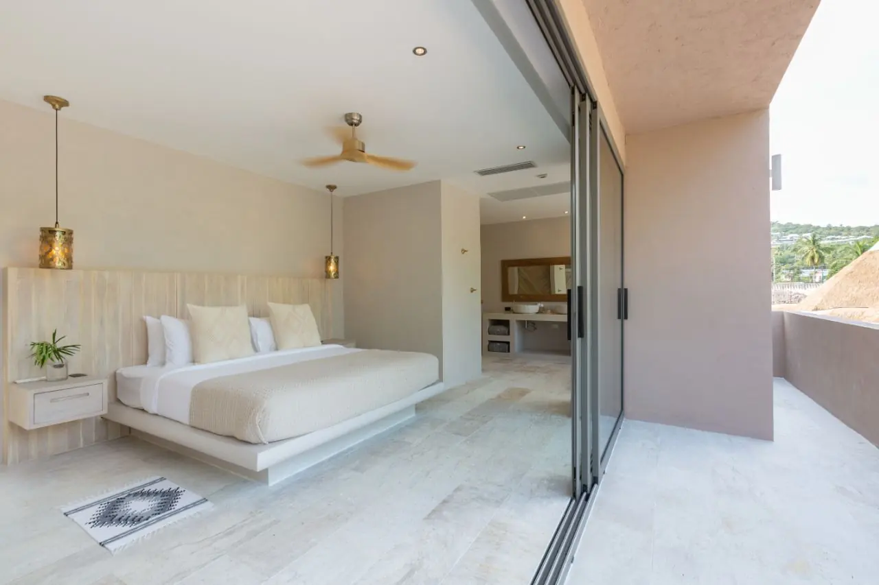 Upstairs bedroom with a king bed and lantern pendants, its full-height glass doors open to a balcony over treetops and a thatched roof.