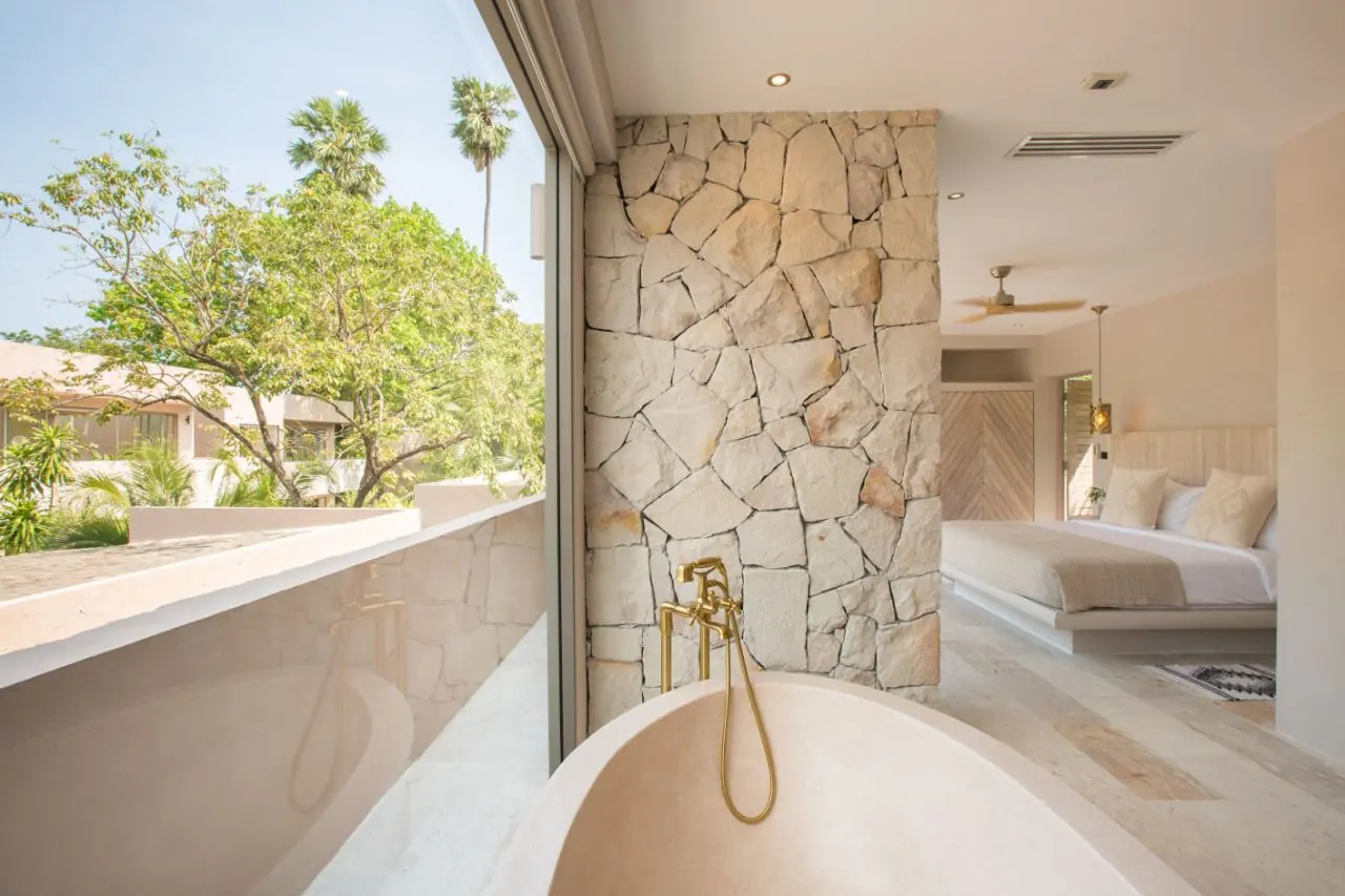 Freestanding stone bathtub with a brass mixer by a full-height window over trees, with a rough stone wall and the bedroom beyond.