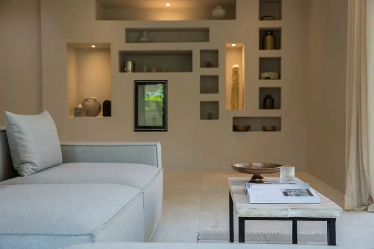 Lounge with a grey sofa and a stone-topped coffee table with a bronze bowl, a glass and a book, before a wall of lit niches holding ceramics.