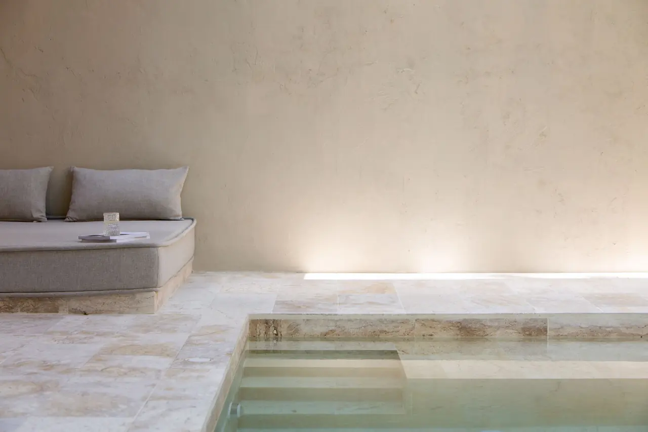 Cushioned grey daybed against a plaster wall with a glass on a magazine, beside the steps of a shallow plunge pool on a travertine deck.