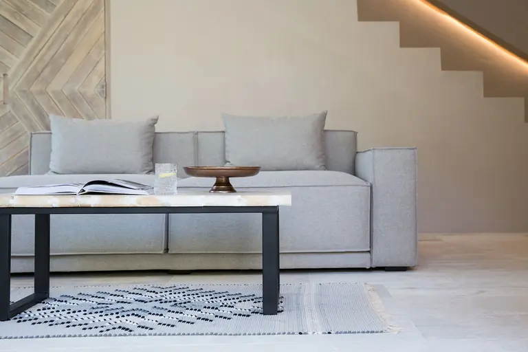 Grey sofa and a black-framed coffee table with a bronze bowl, a glass and an open book on a woven rug, under a staircase with a lit edge beside a chevron timber door.