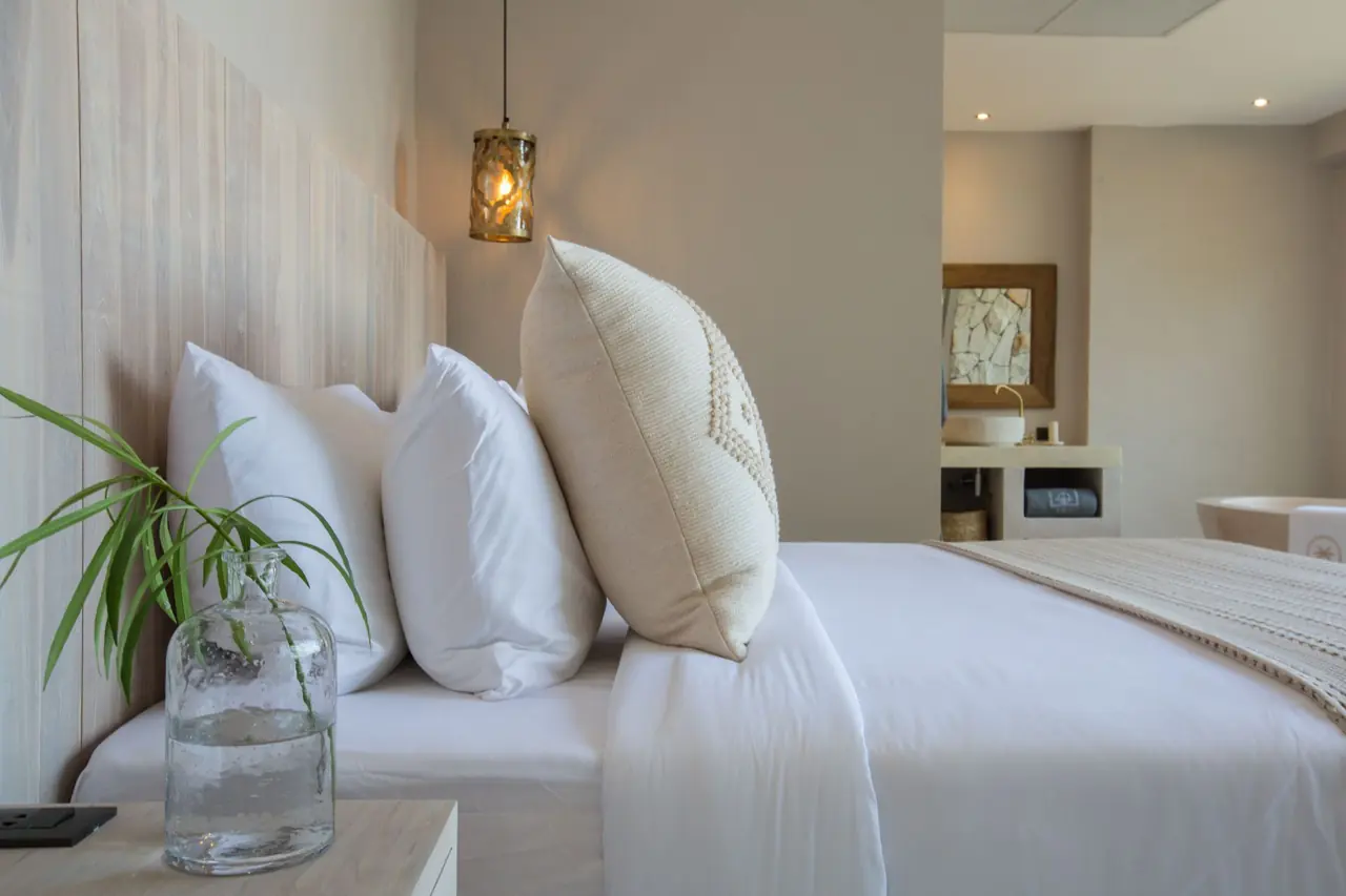 Close view along a made bed with white pillows and a cream cushion, a glass bottle with a green sprig on the nightstand and the vanity beyond.