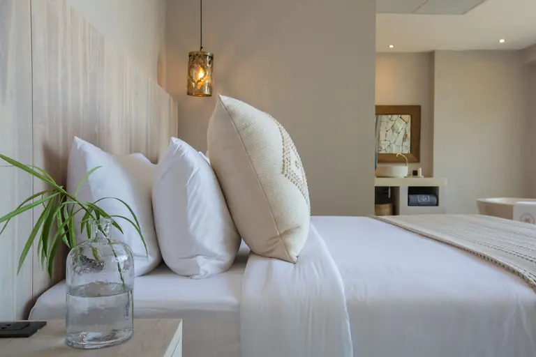 Close view along a made bed with white pillows and a cream cushion, a glass bottle with a green sprig on the nightstand and the vanity beyond.