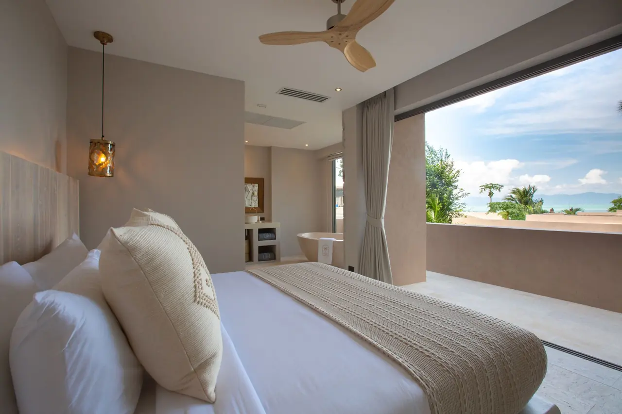 Bedroom with a knitted throw on the bed and a freestanding bathtub behind, opening onto a terrace with a view over rooftops and trees to the sea and distant hills.