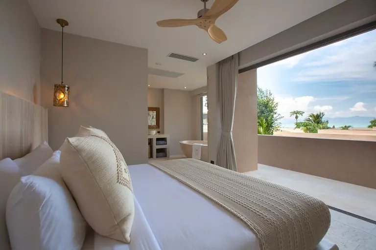 Bedroom with a knitted throw on the bed and a freestanding bathtub behind, opening onto a terrace with a view over rooftops and trees to the sea and distant hills.