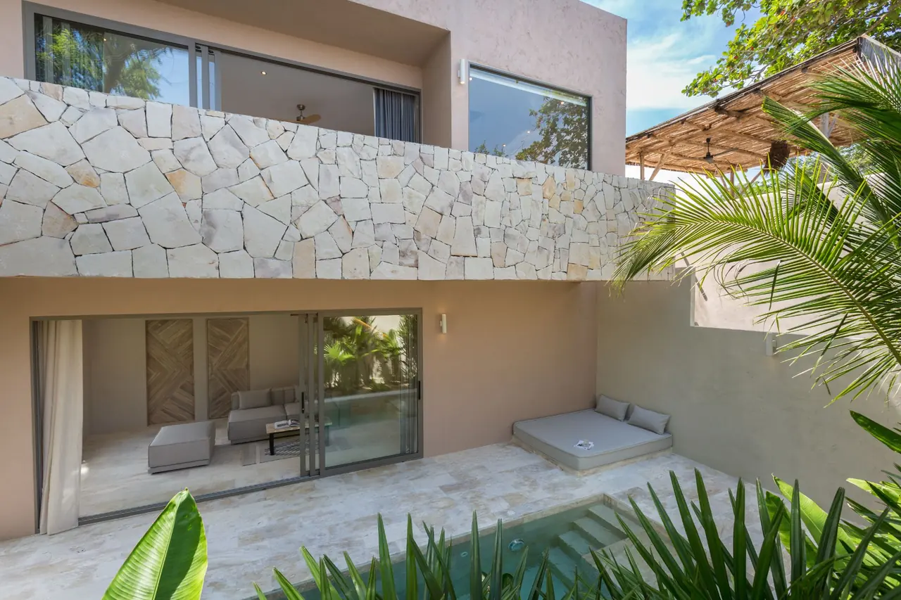 Two-storey suite with a stone-clad upper floor above a living room with open glass doors, a cushioned daybed and the steps of a plunge pool among tropical plants.