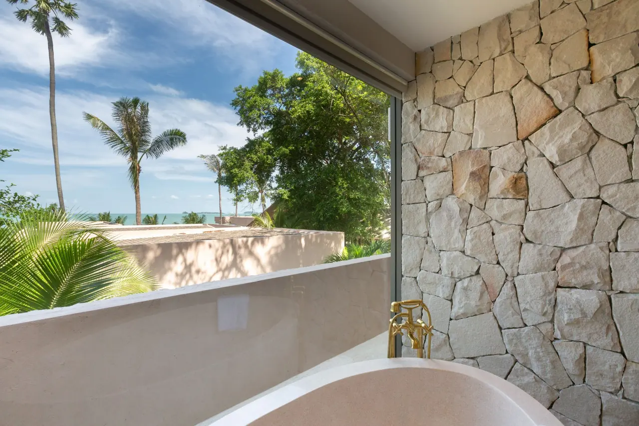 Freestanding stone bathtub with a brass mixer beside a rough stone wall and a large window looking over palms and trees to the sea.