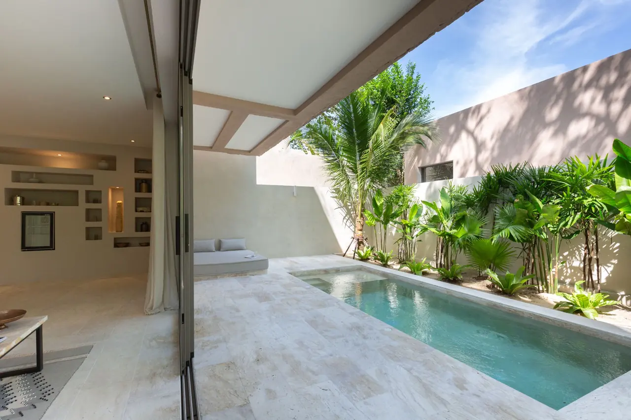 Living room with lit wall niches opening through full-height glass doors onto a narrow private pool lined with palms and tropical plants.