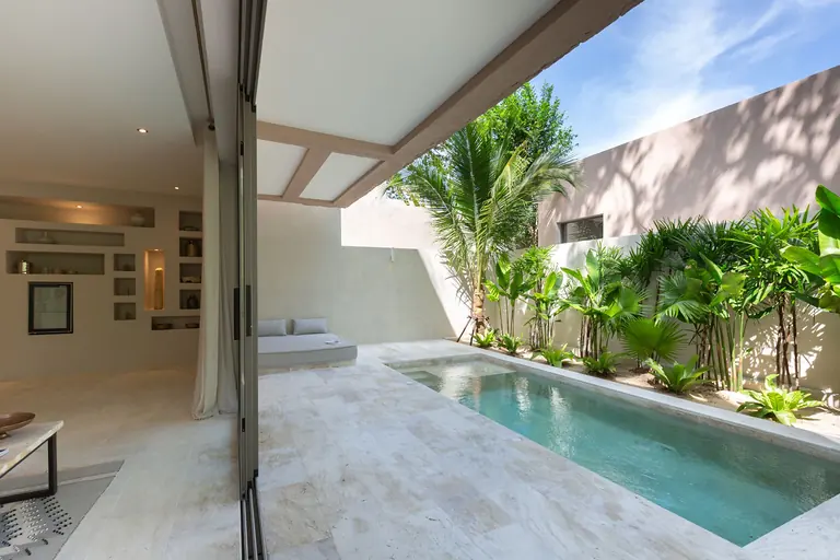 Living room with lit wall niches opening through full-height glass doors onto a narrow private pool lined with palms and tropical plants.