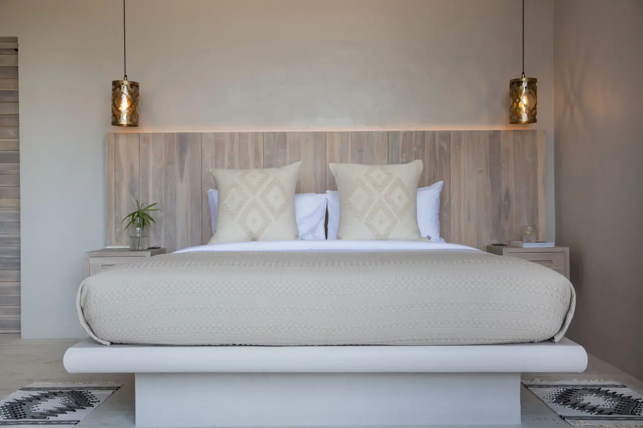 Bed with diamond-pattern cushions against a lit timber headboard between two lantern pendants and floating bedside drawers.