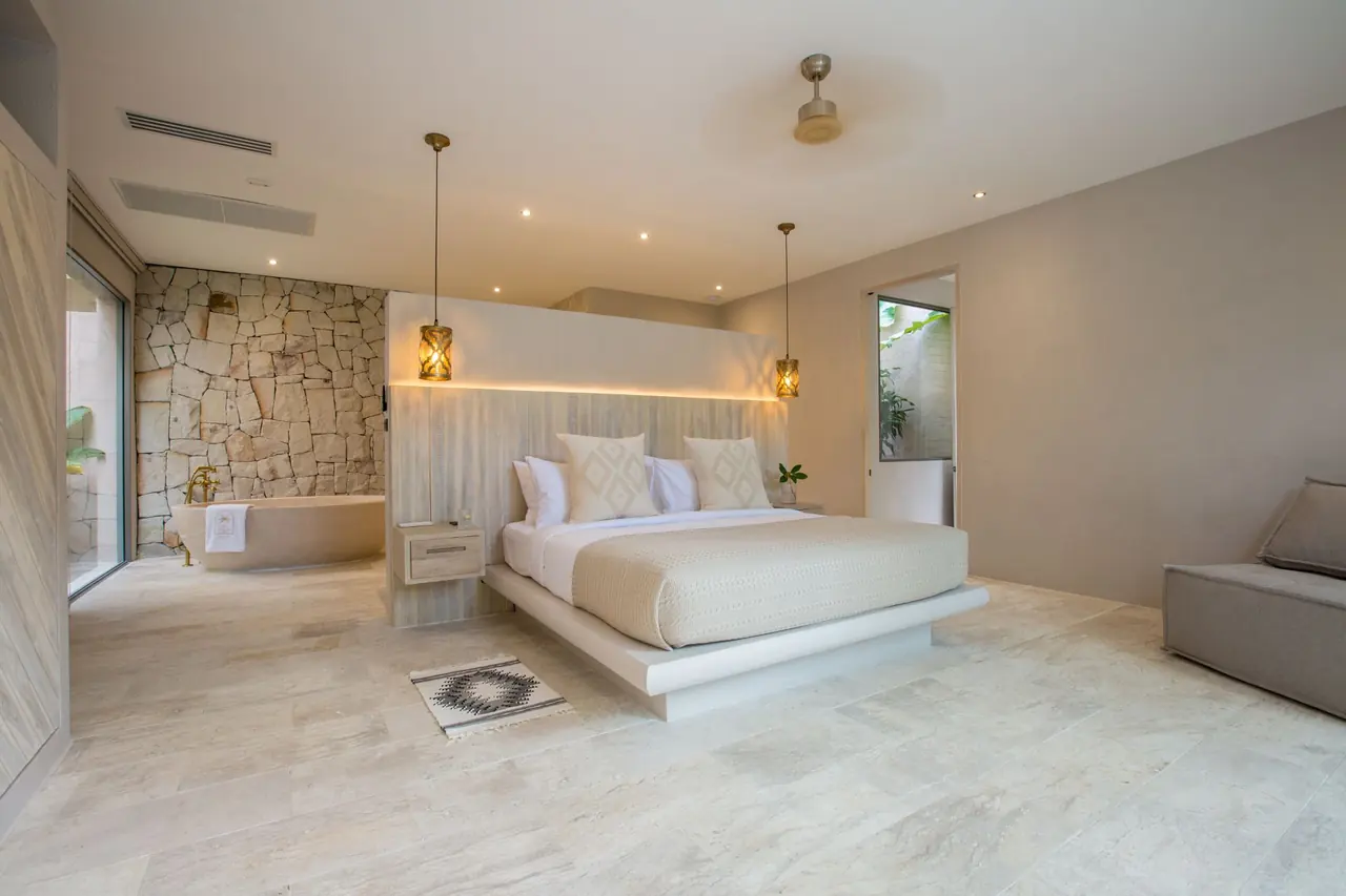 Spacious suite with a bed against a lit timber headboard wall, a freestanding stone bathtub behind it against a rough stone wall, and a grey seat at right.