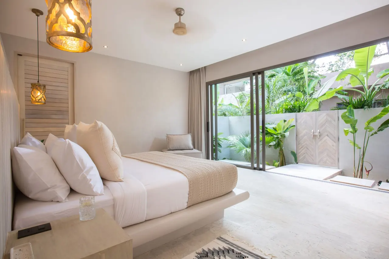 Bed with a glass on the bedside ledge beside a large lantern pendant, facing sliding doors open to a walled garden of banana plants and palms.