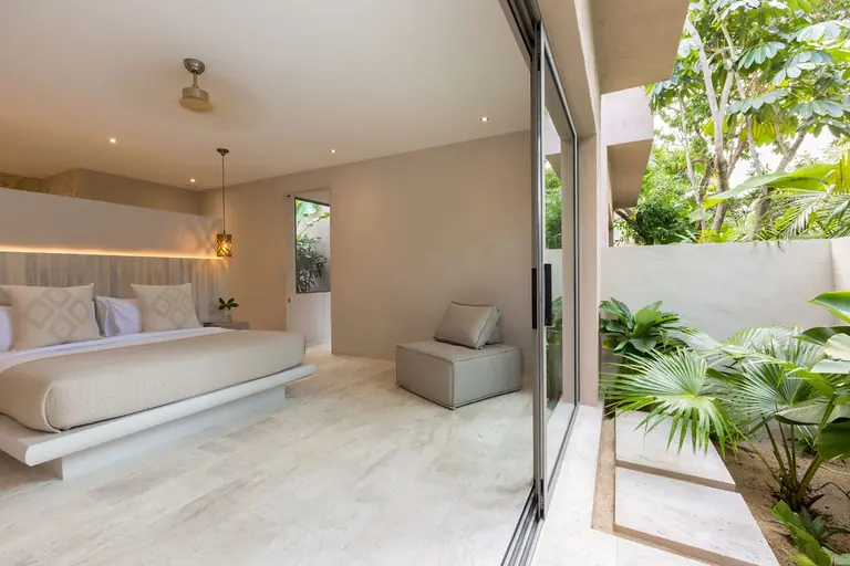 Bedroom with a bed, a lantern pendant and a grey seat, beside a full-height glass door onto a walled garden of tropical plants.