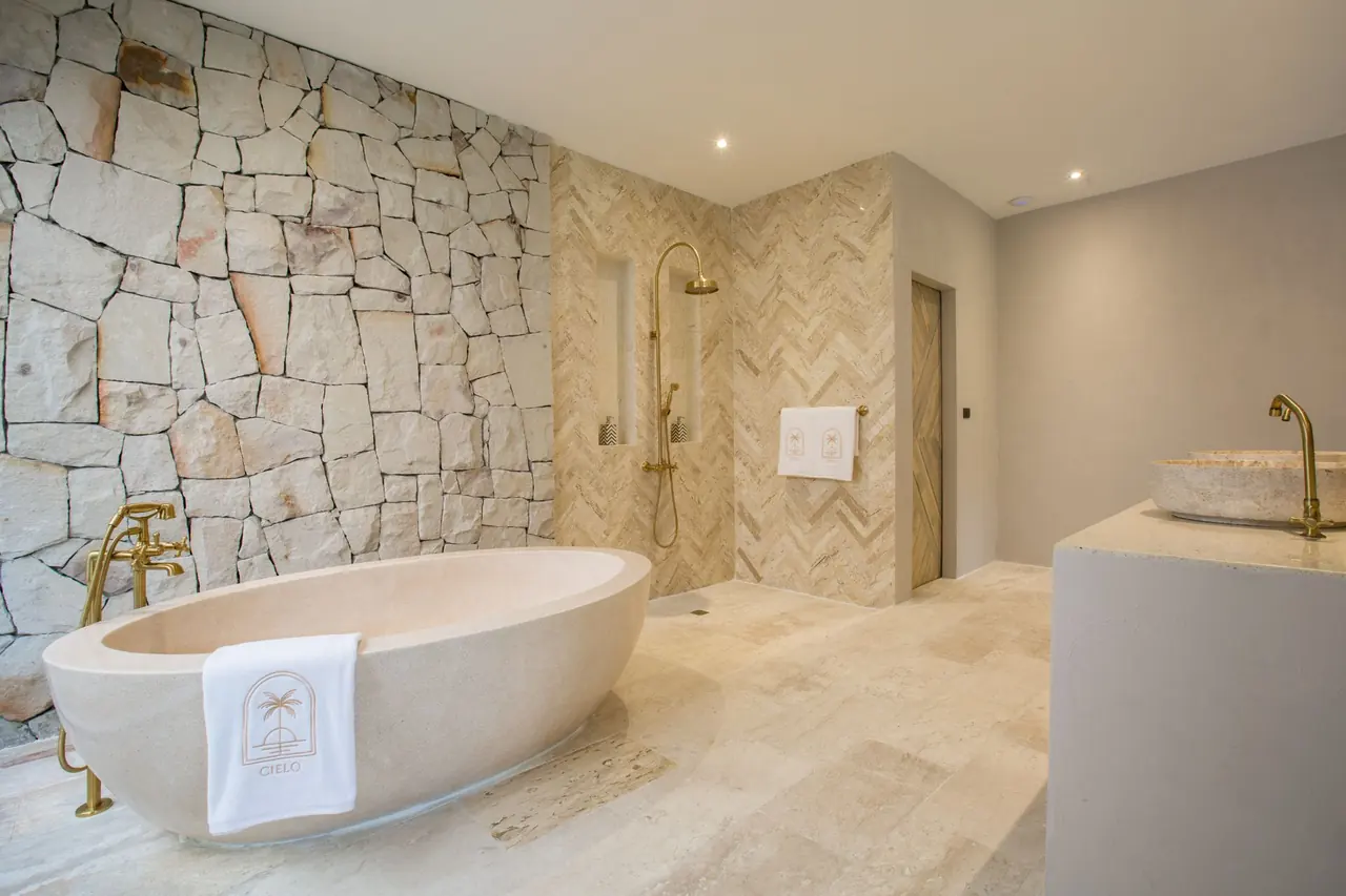 Bathroom with a freestanding stone bathtub and a Cielo logo towel against a stone wall, a brass rain shower on herringbone tiles and a stone basin.