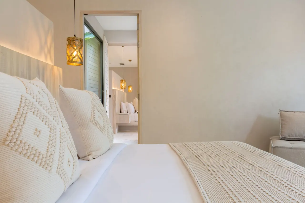 Close view across cushions and a knitted throw to an open door into a second bedroom with its own bed and pendant lamps.