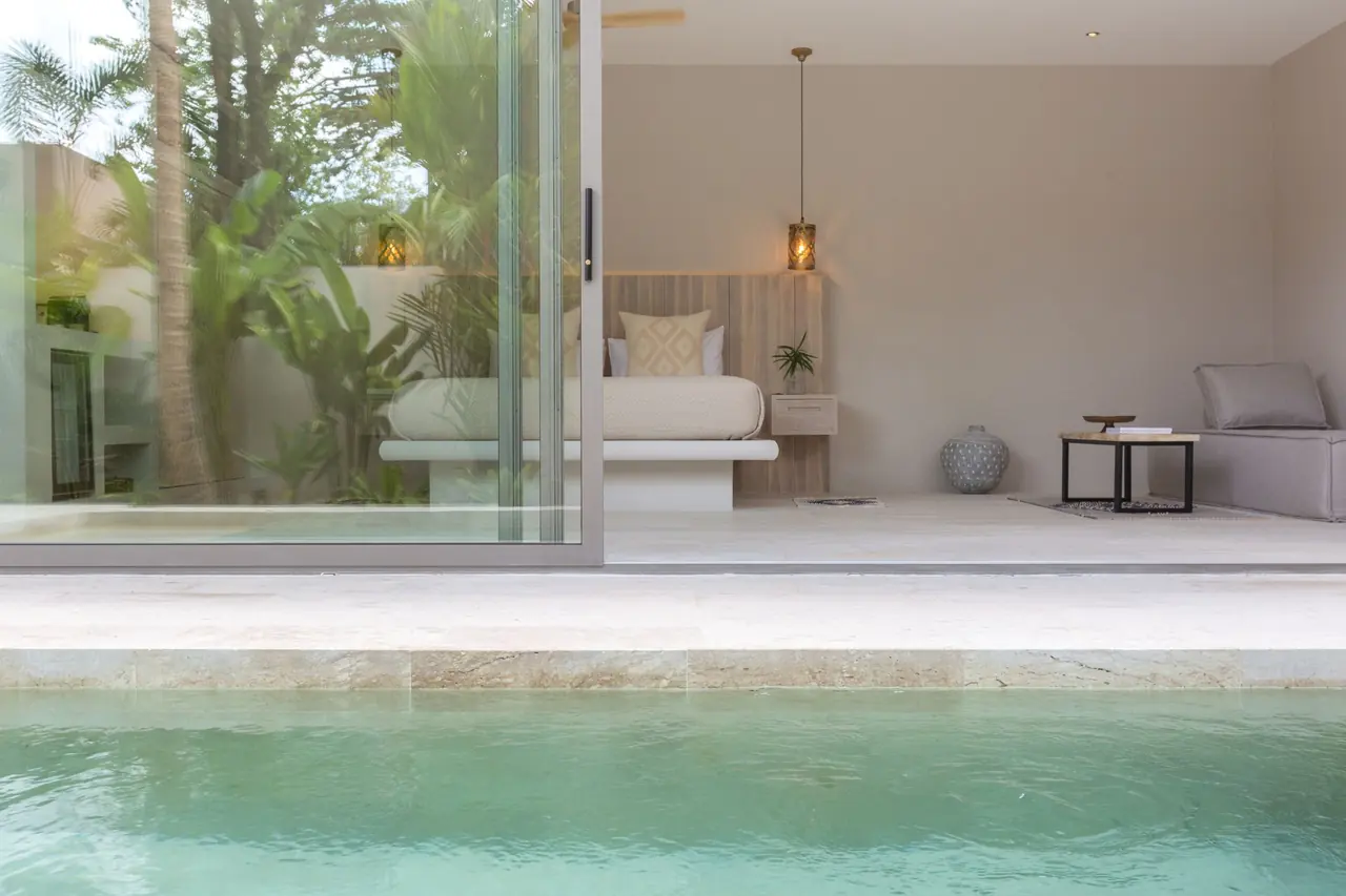 Bedroom seen across a private plunge pool through open glass doors, with the bed, lantern pendants, a side table and a grey daybed.