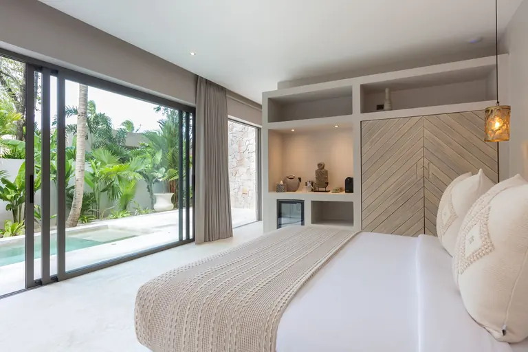 Bed with a knitted throw facing a built-in wardrobe with chevron doors and lit shelves, beside floor-to-ceiling glass doors to the plunge pool and garden.